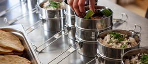   Image showing a line up of Dabba tins, filled with Wasted Kitchen Dabba Feast take away food. A hand is placing a lime wedge into one of the tins. 